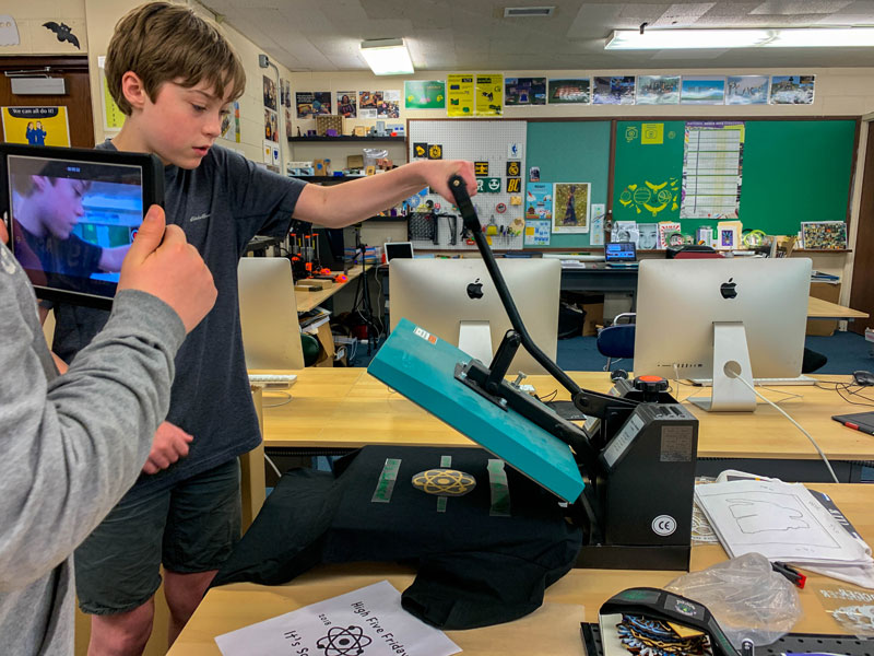 Two 7th grade students use the heat press to adhere vinyl on to their "High Five Friday" shirts created for their science class. One student presses while the other documents the process via time lapse video.